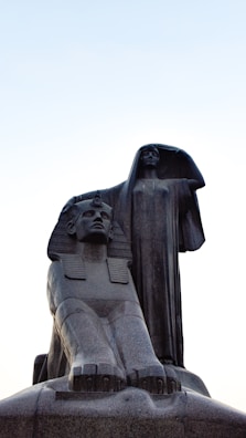 Large stone statues depicting an Egyptian sphinx and a robed woman standing behind it. The sphinx has a stoic expression with traditional Egyptian headgear, while the robed woman stretches one arm up, creating an impression of grace and authority. The background is a clear sky during daytime.