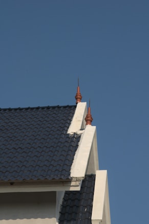A section of a building roof with dark blue tiles and white trim is prominently featured, with the sky as the background. Decorative spires are placed on top of the roof, adding an architectural detail.