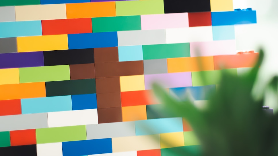 A wall made of colorful interlocking plastic bricks arranged in a pattern, with various shades including red, yellow, blue, green, and black. The foreground features blurred green leaves, adding a touch of nature to the scene.