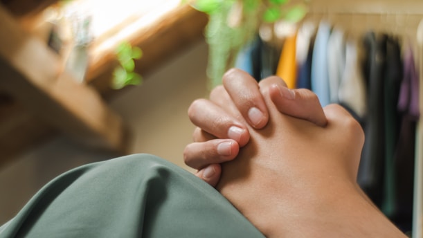 Two hands are clasped together, perhaps in a gesture of prayer or contemplation. In the background, soft-focused elements such as hanging clothes and vibrant green plants suggest a cozy indoor setting with natural light streaming in from a window.