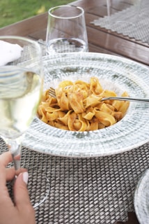 A plate of pasta with a tomato-based sauce is placed on a textured placemat. A fork is resting on the pasta, and a hand is holding a glass of white wine nearby. The table also features two empty water glasses, and a hint of green grass is visible in the background.