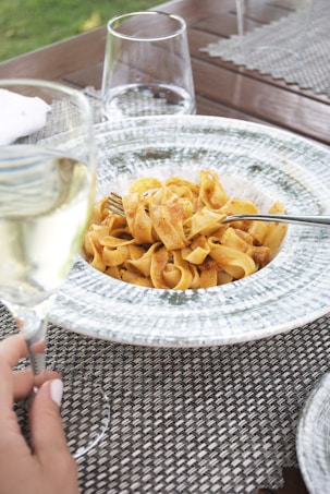 A plate of pasta with a tomato-based sauce is placed on a textured placemat. A fork is resting on the pasta, and a hand is holding a glass of white wine nearby. The table also features two empty water glasses, and a hint of green grass is visible in the background.