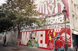 A large mural on a building wall features vibrant graffiti art. In the center, a tribute to Stan Lee with the word 'Marvel' prominently displayed. Colorful characters like Iron Man and Spider-Man are depicted. A tree and a lamp post are in the foreground, adding depth to the scene. Various tags and graffiti adorn the upper part of the wall.