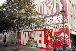 A large mural on a building wall features vibrant graffiti art. In the center, a tribute to Stan Lee with the word 'Marvel' prominently displayed. Colorful characters like Iron Man and Spider-Man are depicted. A tree and a lamp post are in the foreground, adding depth to the scene. Various tags and graffiti adorn the upper part of the wall.