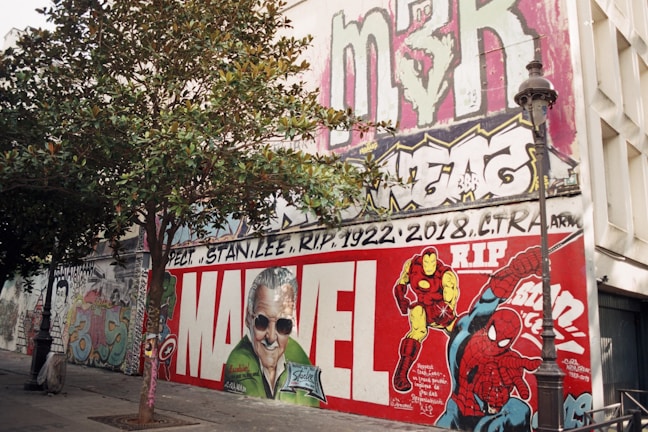 A large mural on a building wall features vibrant graffiti art. In the center, a tribute to Stan Lee with the word 'Marvel' prominently displayed. Colorful characters like Iron Man and Spider-Man are depicted. A tree and a lamp post are in the foreground, adding depth to the scene. Various tags and graffiti adorn the upper part of the wall.