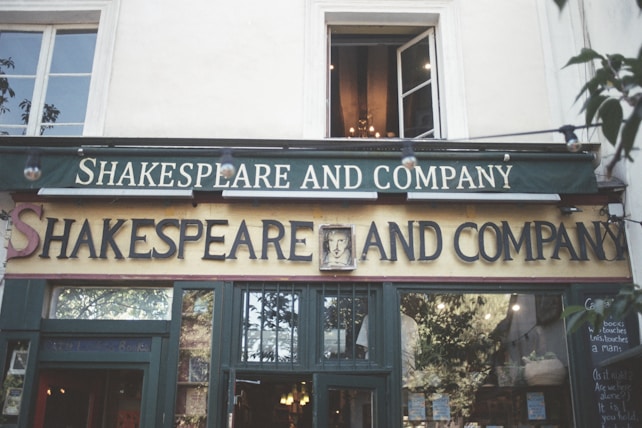The exterior of a bookstore named 'Shakespeare and Company' with old-style signage and an open window above. Green and cream colors dominate the facade, while soft lighting inside can be seen from outside. Light strings hang across the image, contributing to a cozy atmosphere. Plants and foliage partially frame the shop.
