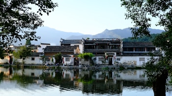 Traditional Chinese architecture is reflected in a serene body of water with green trees framing the scene. The buildings are built with classic white walls and dark, tiled roofs, creating a contrast against the backdrop of distant mountains under a clear sky.