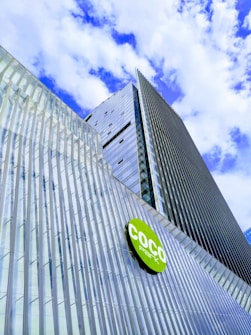 A modern high-rise building with a sleek, reflective glass facade stretches towards a partly cloudy sky. The structure includes a green circular sign with the text 'COCO Park'. The glass panels are arranged in vertical lines, creating a textured and dynamic appearance.