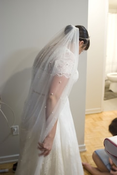 A bride dressed in a white wedding gown with delicate lace detailing and a sheer veil adorned with small pearls is leaning slightly forward. She is standing indoors, near a partially visible bathroom with a tiled floor. Another person, possibly a child, can be seen kneeling in the background.