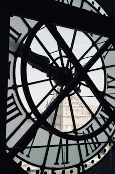 A large clock face with Roman numerals is seen from the inside, providing an intricate view of its inner workings. The background reveals a historic building, likely of European architecture, framed by the clock's transparent sections.