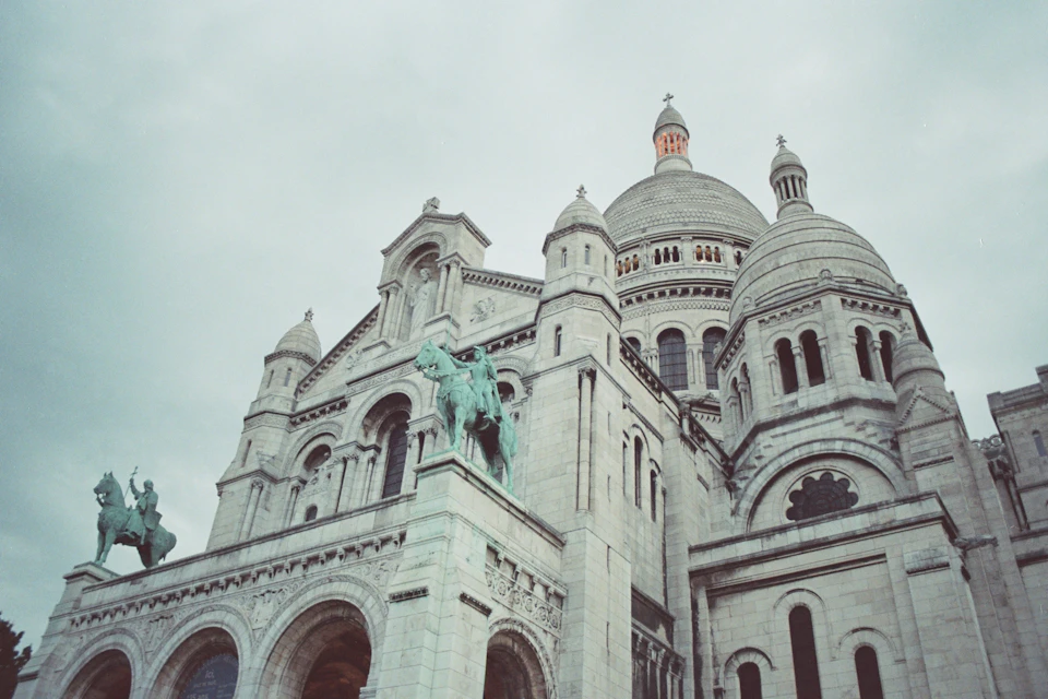 The image depicts a large, ornate white building with multiple domes and towers, standing tall against an overcast sky. The building features intricate architectural details that showcase its grandeur. Atop the structure, two statues can be seen - one of a man on horseback and another of a man on foot.

The perspective of the image is from below, looking up at the imposing stature of the building. The cloudy sky casts a soft light over the scene, highlighting the architectural features of the building while also creating an atmospheric mood.