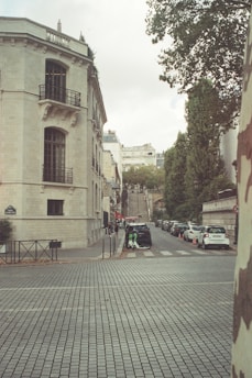 A European city street features a large stone building with wrought iron balconies and detailed architecture. The cobblestone pavement leads up to a set of stairs in the distance. Several parked cars line the street, and a few pedestrians are visible. The trees and parked scooters add to the urban scenery, and the sky is overcast.