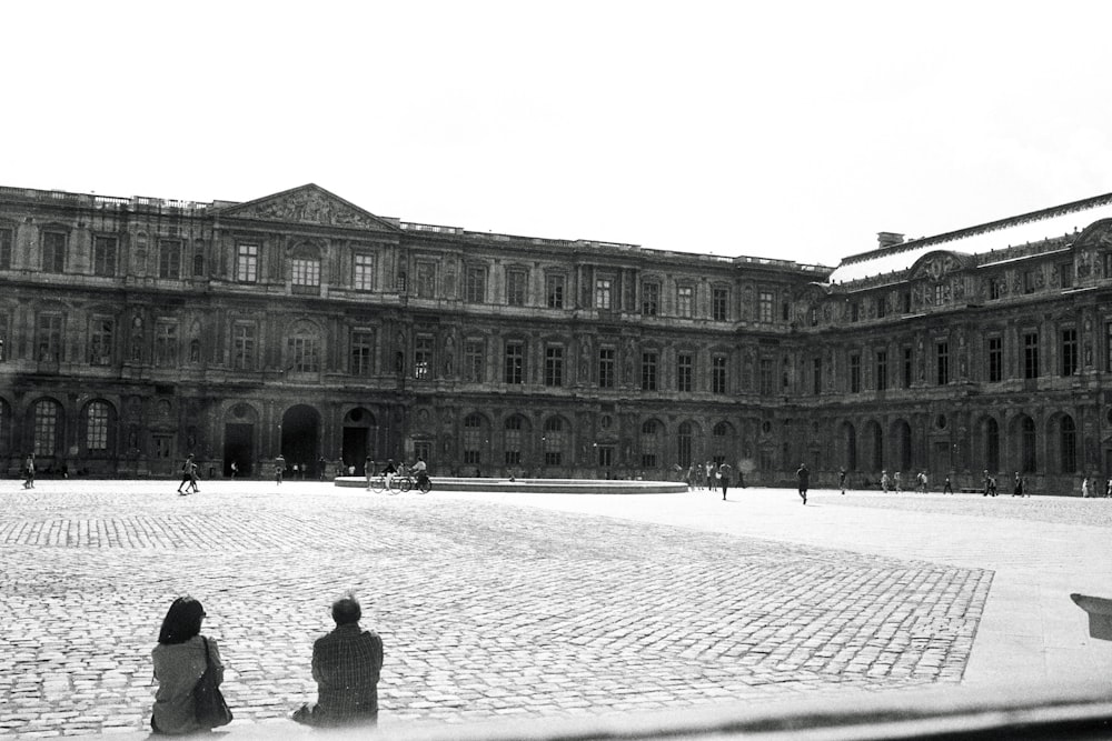 two people sitting on the ground in front of a large building