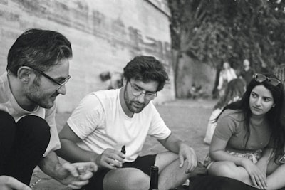 Three people are sitting together outdoors, engaged in a friendly conversation. The scene is captured in a candid moment, with a relaxed atmosphere. They are casually dressed and appear to be enjoying each other's company.