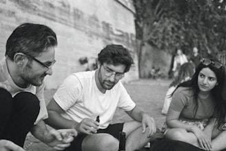 Three people are sitting together outdoors, engaged in a friendly conversation. The scene is captured in a candid moment, with a relaxed atmosphere. They are casually dressed and appear to be enjoying each other's company.
