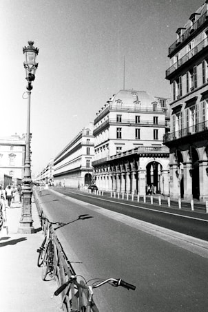 A black-and-white photograph of a long, elegant row of historic buildings with traditional architecture. A wide, empty street runs alongside, with a bicycle leaning against a fence in the foreground. A classic street lamp stands prominently on the left. People are walking along the pavement, providing a sense of bustling city life.