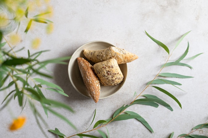 A ceramic plate holds three different types of bread with seeds and grains on a light gray surface. Surrounding the plate, there are delicate green leaves and yellow flowers, adding a natural and fresh ambiance.