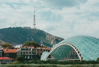 A modern glass-arched bridge stretches gracefully across a river with a backdrop of a lush, green hillside. The hill has a prominent telecommunications tower at the peak. In the foreground, there are several low-rise buildings with colorful roofs and trees scattered around.