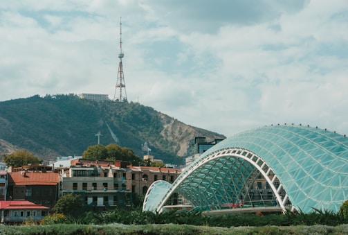 A modern glass-arched bridge stretches gracefully across a river with a backdrop of a lush, green hillside. The hill has a prominent telecommunications tower at the peak. In the foreground, there are several low-rise buildings with colorful roofs and trees scattered around.