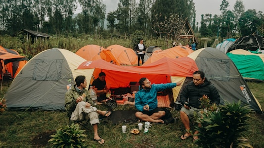 Several tents are set up in a grassy area surrounded by trees, with people sitting and interacting nearby. The setting appears to be a camping site with vibrant tents of different colors. One person is sitting on the ground while others are in folding chairs, engaging in conversation and holding food and drinks.