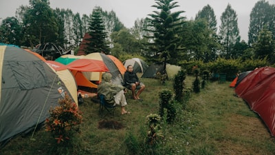 Several colorful tents are set up on a grassy area surrounded by trees. Two people are sitting on camping chairs near the tents, engaged in conversation. The scene is set amidst lush greenery, with tall trees and shrubs providing a natural backdrop.
