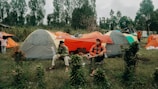 Several tents are set up in a grassy area surrounded by trees, with two people sitting and conversing in front of them. One person is seated casually on a camping chair, and the tents are in shades of gray and orange.
