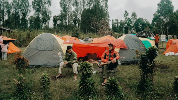 Several tents are set up in a grassy area surrounded by trees, with two people sitting and conversing in front of them. One person is seated casually on a camping chair, and the tents are in shades of gray and orange.