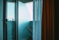 A serene view of a small balcony area is seen through an open glass door, with a modern wire-frame chair positioned against a turquoise wall. Sheer white and mustard yellow curtains drape to the side, allowing a soft, ambient light to fill the space, creating a peaceful atmosphere.