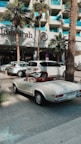 A classic white convertible Mercedes-Benz is parked on a paved driveway in front of a modern hotel with a sign reading 'Jumeirah'. The background features palm trees and a glass building facade, with other parked cars visible nearby.