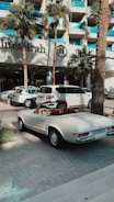 A classic white convertible Mercedes-Benz is parked on a paved driveway in front of a modern hotel with a sign reading 'Jumeirah'. The background features palm trees and a glass building facade, with other parked cars visible nearby.
