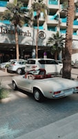 A classic white convertible Mercedes-Benz is parked on a paved driveway in front of a modern hotel with a sign reading 'Jumeirah'. The background features palm trees and a glass building facade, with other parked cars visible nearby.