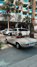 A classic white convertible Mercedes-Benz is parked on a paved driveway in front of a modern hotel with a sign reading 'Jumeirah'. The background features palm trees and a glass building facade, with other parked cars visible nearby.