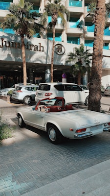 A classic white convertible Mercedes-Benz is parked on a paved driveway in front of a modern hotel with a sign reading 'Jumeirah'. The background features palm trees and a glass building facade, with other parked cars visible nearby.