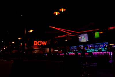 A dimly lit bowling alley interior with neon red 'BOW' signage prominently displayed. The scene includes a row of hanging lights and a bar area illuminated by bright blue and purple lighting. Television screens can be seen above the bar.