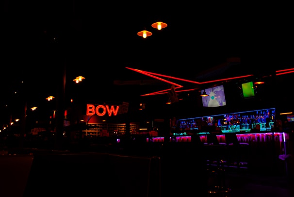A dimly lit bowling alley interior with neon red 'BOW' signage prominently displayed. The scene includes a row of hanging lights and a bar area illuminated by bright blue and purple lighting. Television screens can be seen above the bar.
