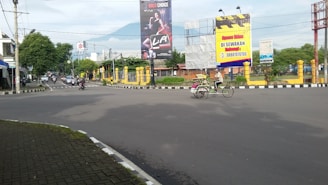 An intersection with a few vehicles such as motorcycles and a becak (pedicab) on the road. Two large billboards are visible, one advertising a beverage and the other offering space for rent. Trees line the street on both sides, and a mountain can be seen in the background under a partly cloudy sky.