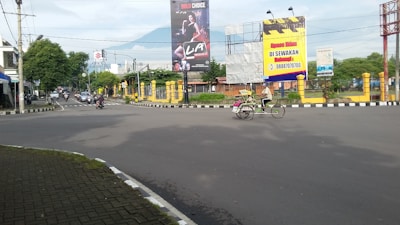 An intersection with a few vehicles such as motorcycles and a becak (pedicab) on the road. Two large billboards are visible, one advertising a beverage and the other offering space for rent. Trees line the street on both sides, and a mountain can be seen in the background under a partly cloudy sky.