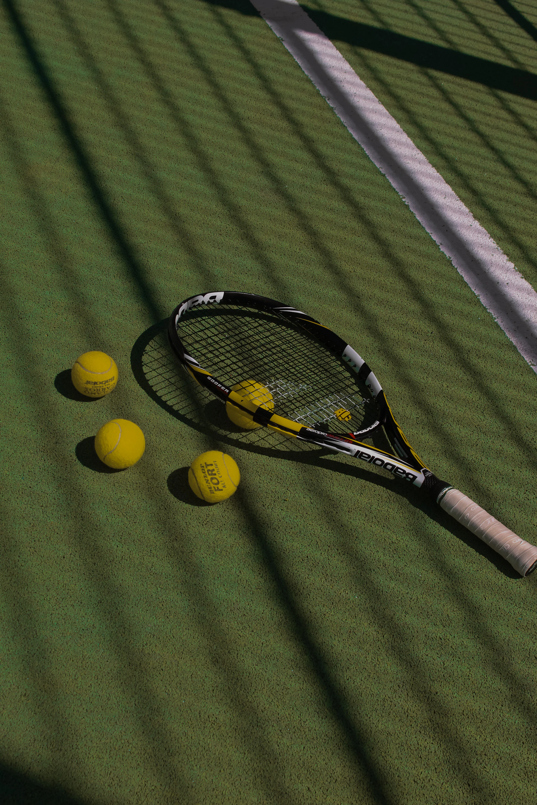 A racquet and three balls lying on a green court in the striped shadow of a fence