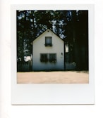 A quaint, small wooden house with a gable roof and white siding. Two rectangular windows at the bottom with flower boxes beneath them, and a smaller window above with its own box. The house is in front of a picket fence and surrounded by tall trees, creating a serene and rustic atmosphere.