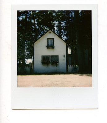 A quaint, small wooden house with a gable roof and white siding. Two rectangular windows at the bottom with flower boxes beneath them, and a smaller window above with its own box. The house is in front of a picket fence and surrounded by tall trees, creating a serene and rustic atmosphere.