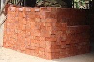 A large stack of red bricks is neatly arranged, forming a rectangular structure. Wooden beams and parts of a building framework are visible in the background. The ground is sandy, indicating an unfinished or construction setting.
