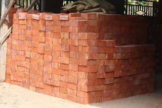 A large stack of red bricks is neatly arranged, forming a rectangular structure. Wooden beams and parts of a building framework are visible in the background. The ground is sandy, indicating an unfinished or construction setting.