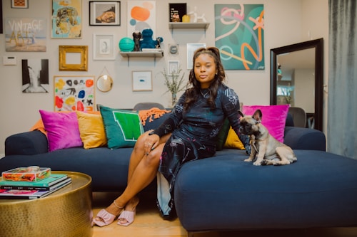 A woman is sitting on a modern dark blue couch surrounded by vibrant pillows in shades of pink, yellow, and green. Beside her sits a small dog with pointed ears. The room is decorated with an eclectic collection of artwork and frames on the wall, creating a stylish and cozy atmosphere. A round, golden coffee table with books is in the foreground.