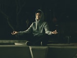 A person is meditating outdoors while sitting crossed-legged on a concrete platform. They are wearing a hooded sweatshirt and appear relaxed with their eyes closed and hands in a meditative mudra. The setting is dimly lit, creating a peaceful and introspective atmosphere.
