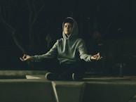 A person is meditating outdoors while sitting crossed-legged on a concrete platform. They are wearing a hooded sweatshirt and appear relaxed with their eyes closed and hands in a meditative mudra. The setting is dimly lit, creating a peaceful and introspective atmosphere.