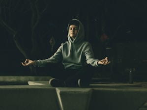 A person is meditating outdoors while sitting crossed-legged on a concrete platform. They are wearing a hooded sweatshirt and appear relaxed with their eyes closed and hands in a meditative mudra. The setting is dimly lit, creating a peaceful and introspective atmosphere.