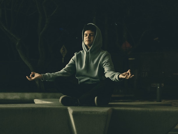 A person is meditating outdoors while sitting crossed-legged on a concrete platform. They are wearing a hooded sweatshirt and appear relaxed with their eyes closed and hands in a meditative mudra. The setting is dimly lit, creating a peaceful and introspective atmosphere.