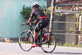 A cyclist in black sportswear rides a black bicycle down a paved road. The rider wears a white helmet and bright red cycling shoes. The surrounding area includes a turquoise wall, some greenery, and part of a building with a tiled roof.