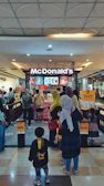 A busy McDonald's outlet in a shopping mall with several people forming a queue at the counter. The entrance and exit are marked with signs in Indonesian. Various digital menu boards displaying food items can be seen behind the counter, and the area is well-lit with a patterned ceiling and shiny floor tiles.