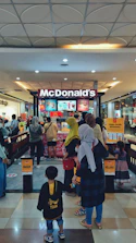 A busy McDonald's outlet in a shopping mall with several people forming a queue at the counter. The entrance and exit are marked with signs in Indonesian. Various digital menu boards displaying food items can be seen behind the counter, and the area is well-lit with a patterned ceiling and shiny floor tiles.